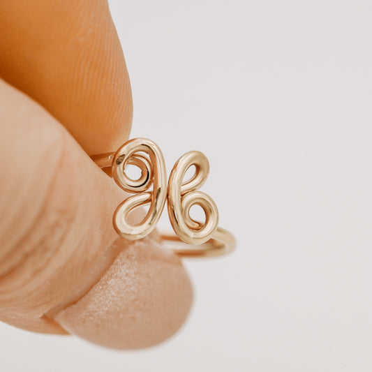 Handmade adjustable wire ring featuring a lowercase cursive letter X in gold on a person's hand with red tipped nails.