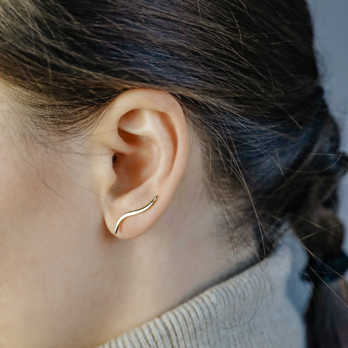 Gold wave ear climber worn across the lower ear, showing how the curved earring rises naturally along the lobe after being inserted through one standard piercing.