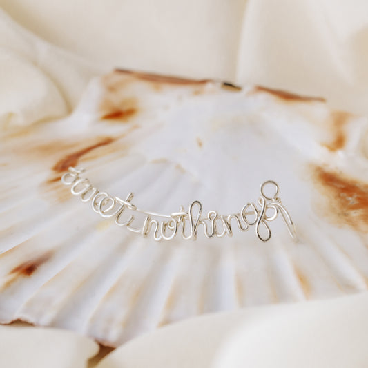Personalized silver name ear climber displayed on a natural scallop shell over soft ivory fabric, featuring custom handwritten wire lettering.