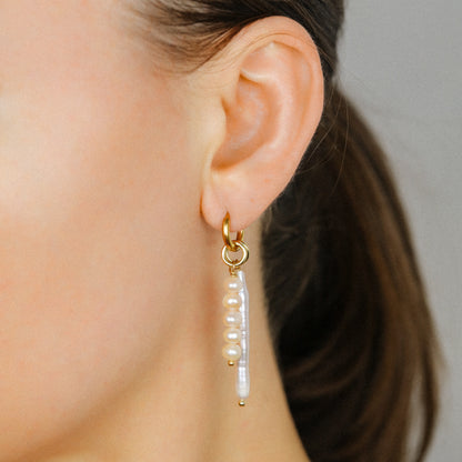 Close-up of a woman wearing an 18k gold plated huggie hoop earring adorned with a long pink baroque stick pearl and a matching five-pearl cluster charm.