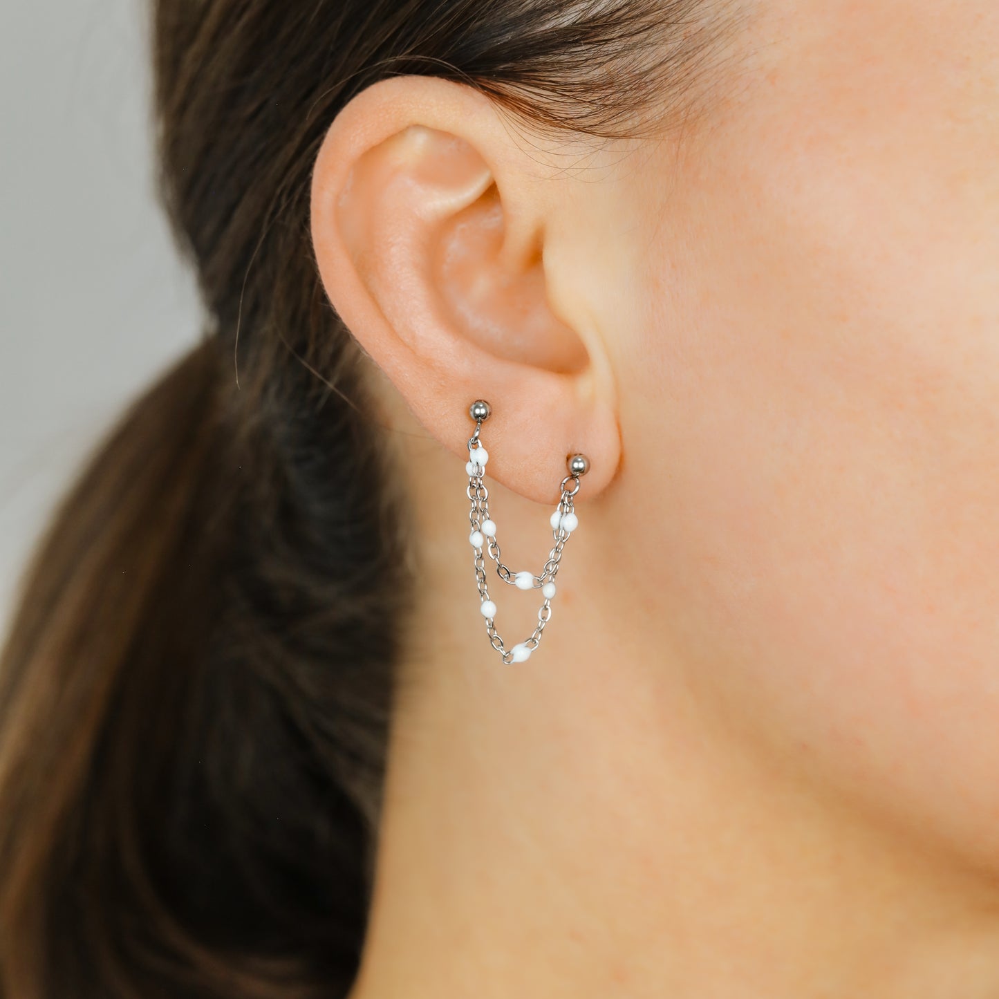 Silver double chain stud earring worn on two lobe piercings with white enamel beads