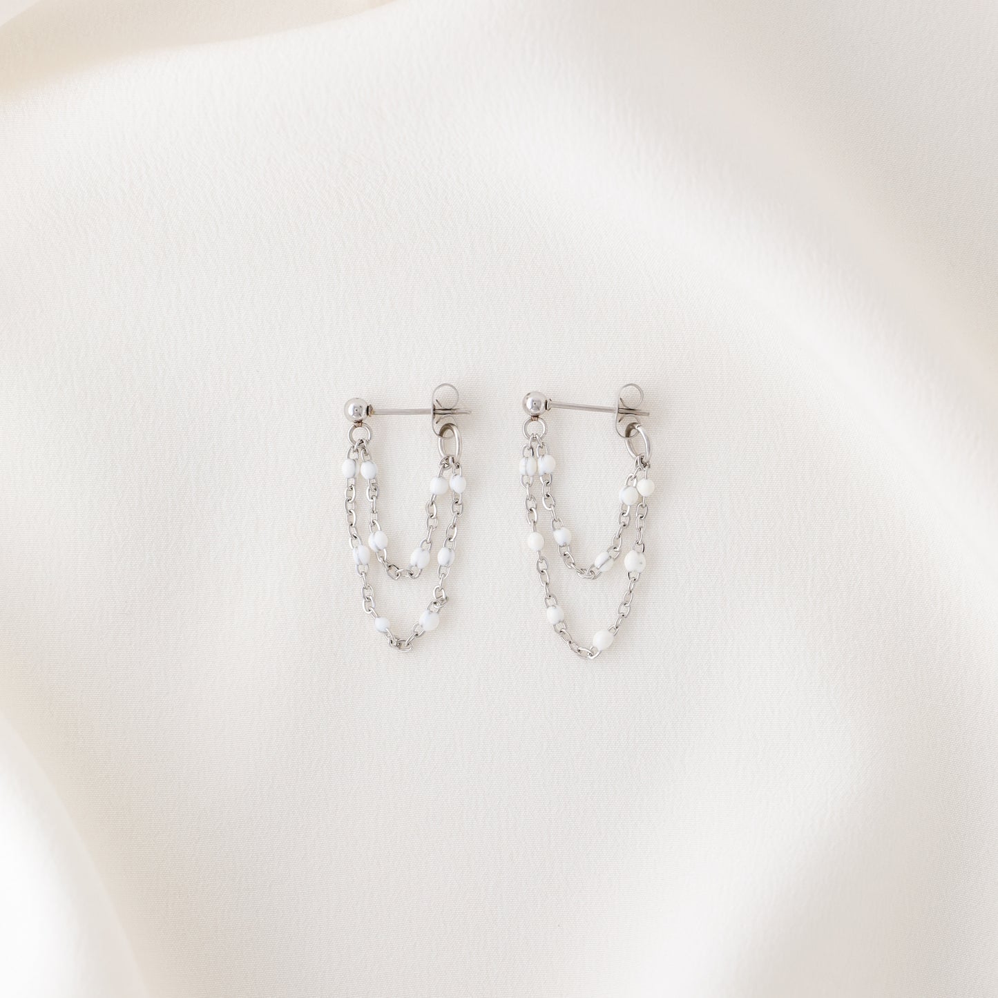 Silver double chain dangle earrings with white enamel bead details displayed on fabric