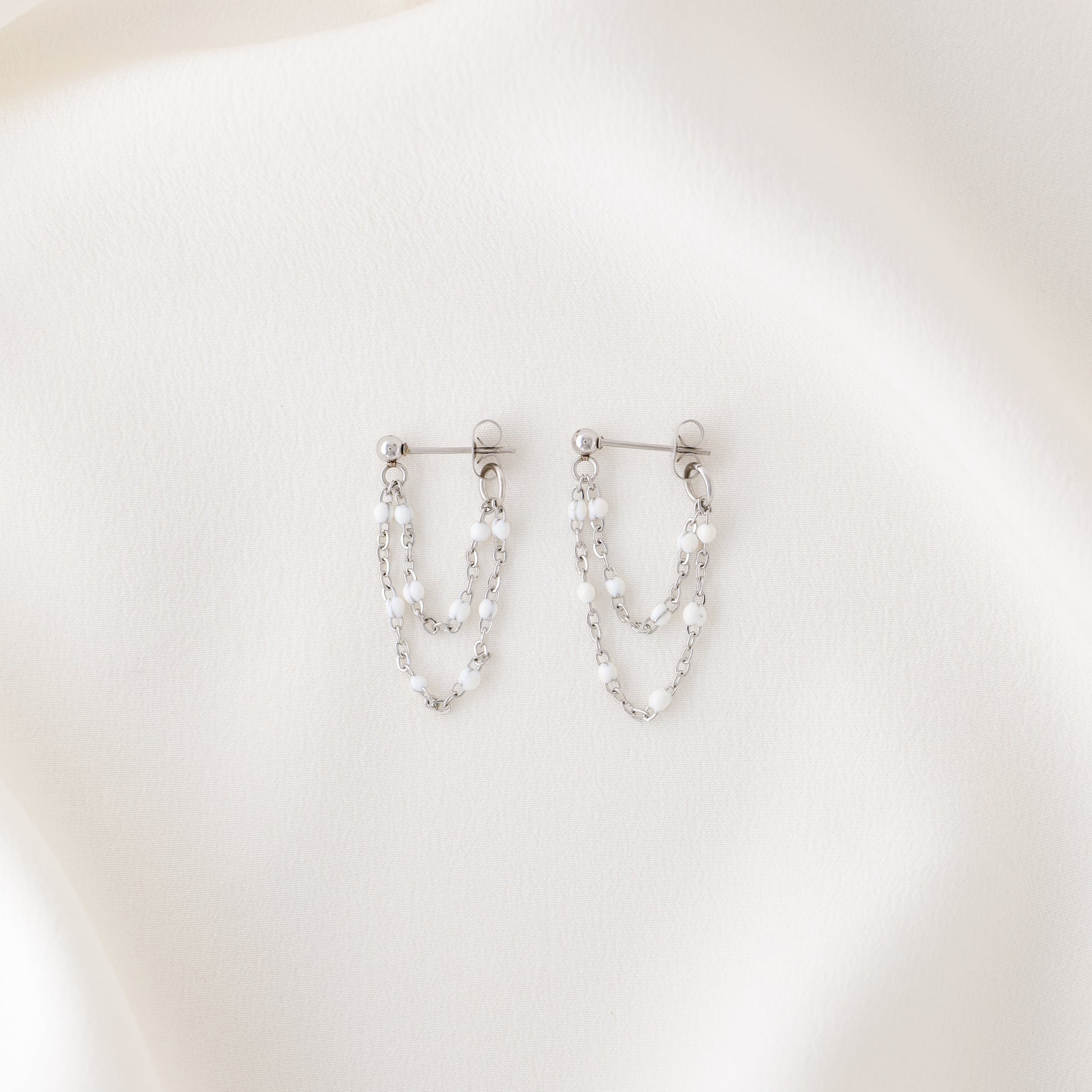 Silver double chain dangle earrings with white enamel bead details displayed on fabric