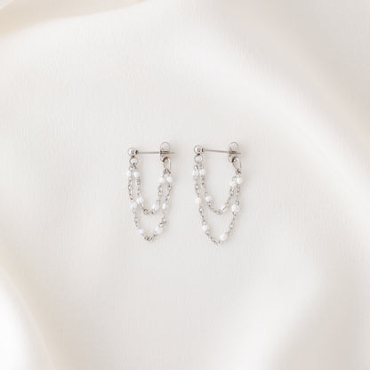 Silver double chain dangle earrings with white enamel bead details displayed on fabric