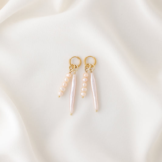 Two gold plated huggie hoop earrings with asymmetrical pink baroque pearl charms resting elegantly on a soft cream-colored silk fabric.