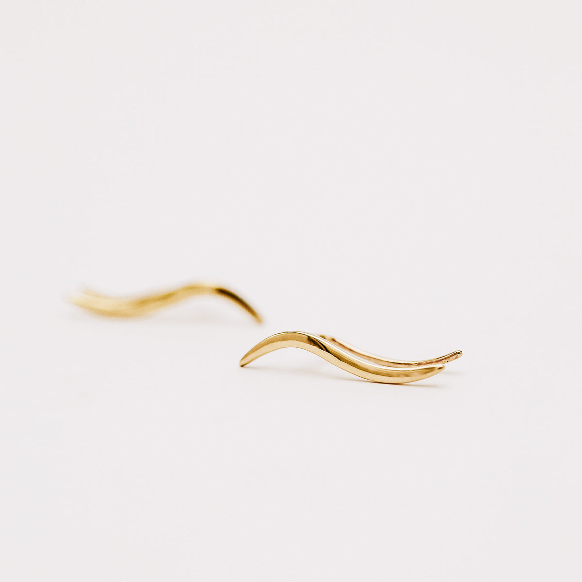 Pair of gold plated wave ear climbers displayed in soft natural light, featuring a flowing curved design with a sleek polished surface.