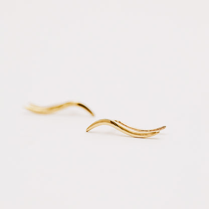Pair of gold plated wave ear climbers displayed in soft natural light, featuring a flowing curved design with a sleek polished surface.