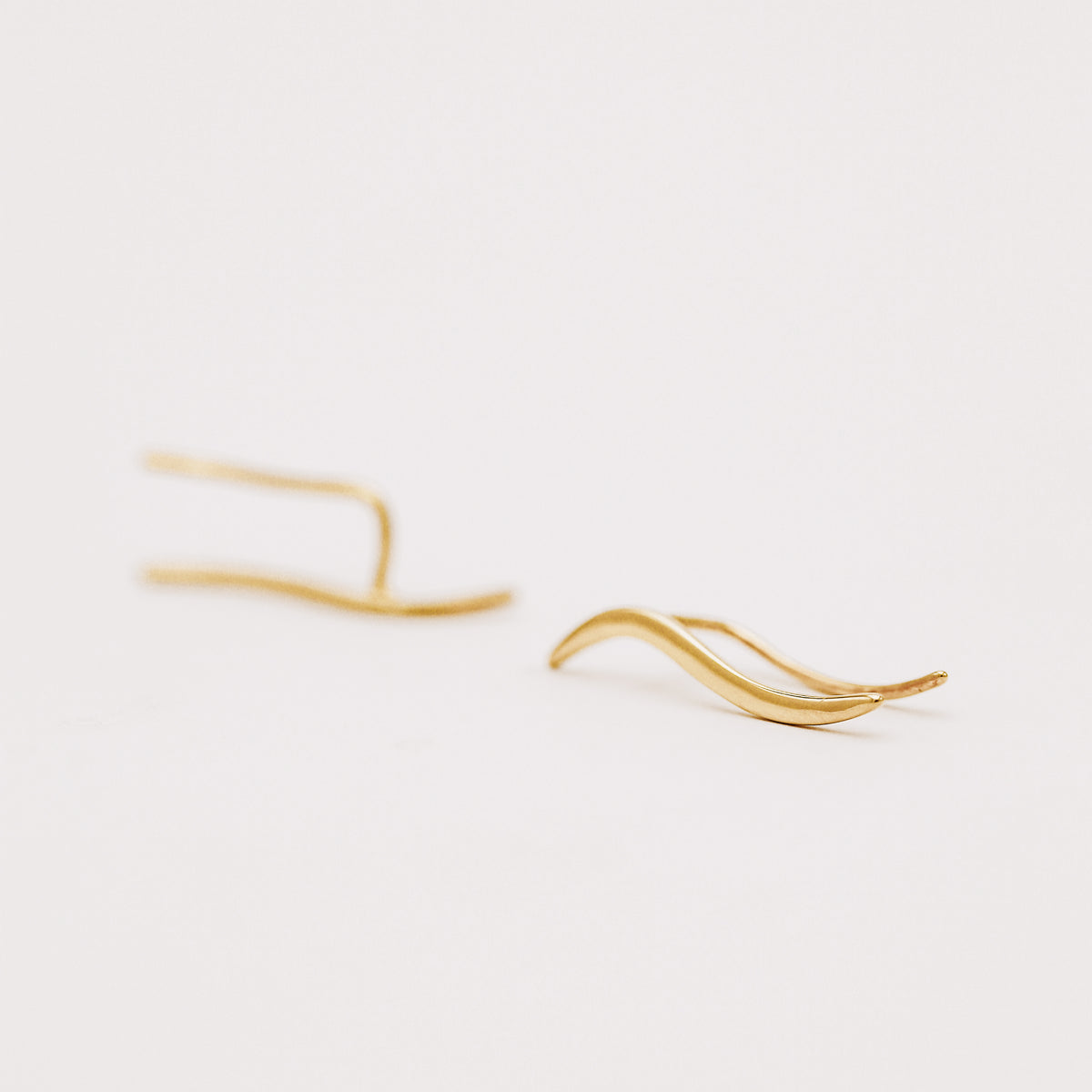 Pair of gold plated wave ear climbers displayed in soft natural light, featuring a flowing curved design with a sleek polished surface.
