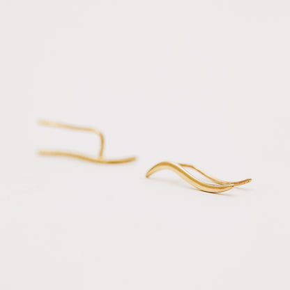 Pair of gold plated wave ear climbers displayed in soft natural light, featuring a flowing curved design with a sleek polished surface.