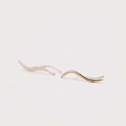 Matching left and right rhodium plated wave ear climbers displayed on a neutral stone surface, showing the mirrored curved silhouettes and high-polish finish.