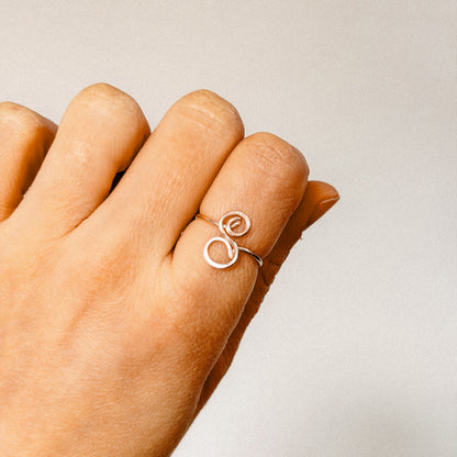 Close-up of a handcrafted gold wire ring with an uppercase cursive letter S, worn on a finger.