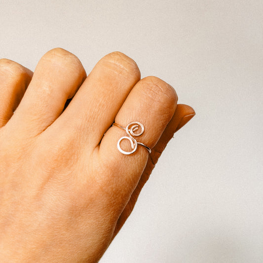 Close-up of a handcrafted gold wire ring with an uppercase cursive letter S, worn on a finger.