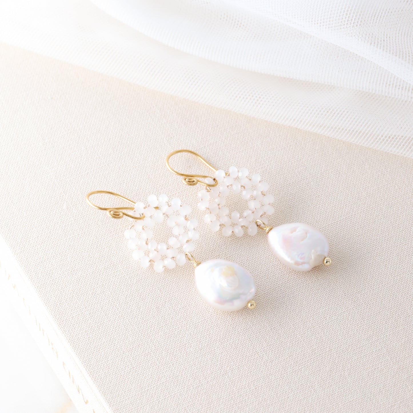 A pair of gold-plated baroque freshwater pearl earrings with woven glass crystal wreaths resting on a textured cream-colored book.