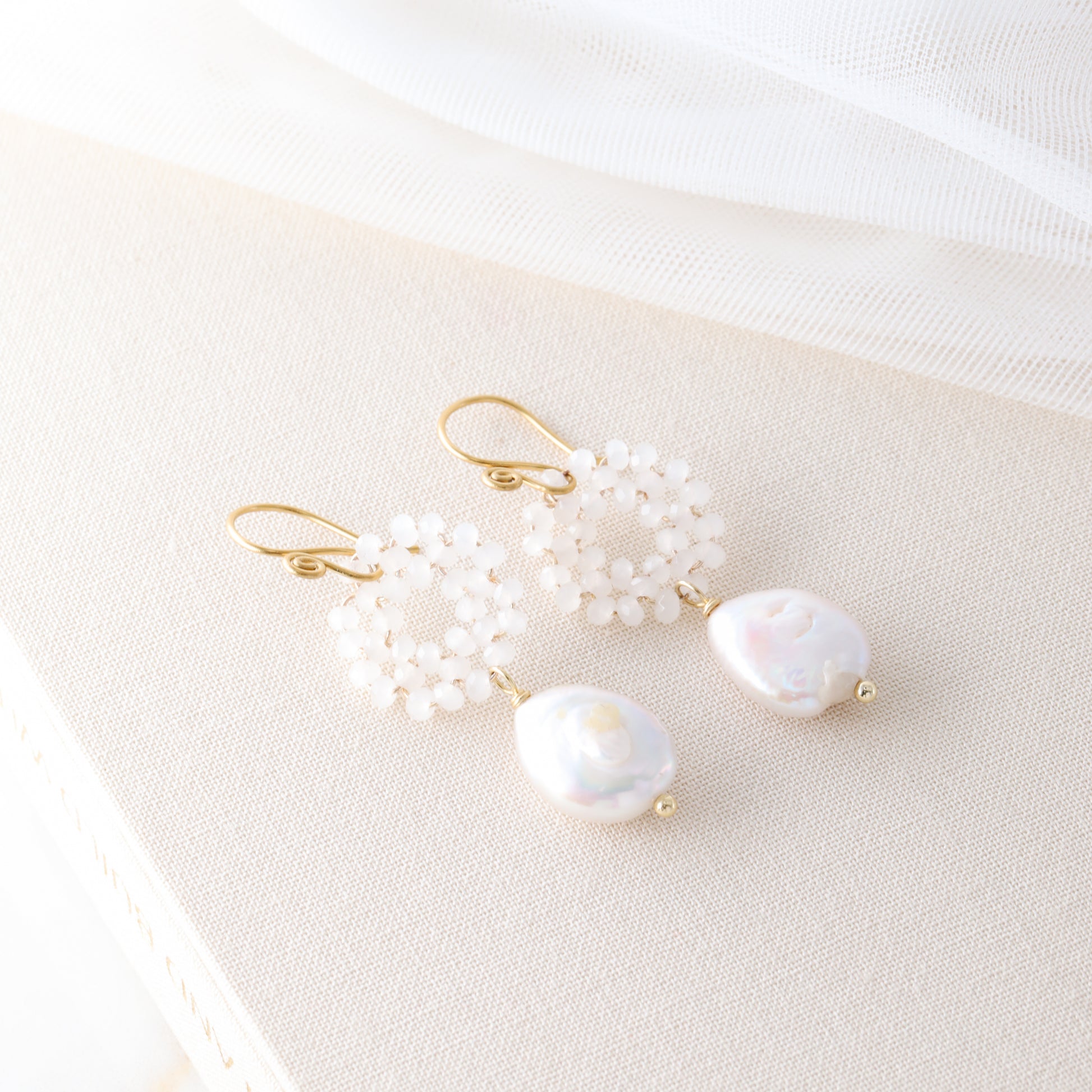 A pair of gold-plated baroque freshwater pearl earrings with woven glass crystal wreaths resting on a textured cream-colored book.