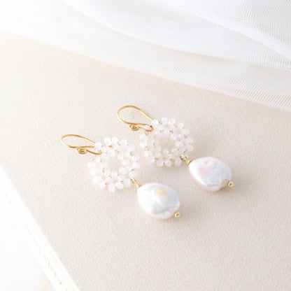 A pair of gold-plated baroque freshwater pearl earrings with woven glass crystal wreaths resting on a textured cream-colored book.