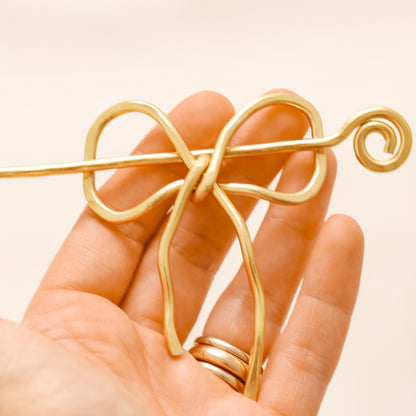 Handmade aluminum bow shawl pin brooch with matching removable stick pin, shown in gold and available in silver or gold finish, designed as a decorative and functional scarf fastener for shawls, sweaters, cardigans, knit wraps and crochet pieces, with a romantic bow shape and minimalist metal design.
