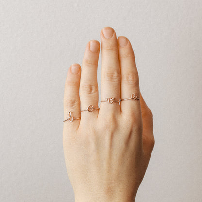 Close-up of a handmade 14k rose gold filled wire ring shaped into the letter L, O, V and E on a woman's fingers.