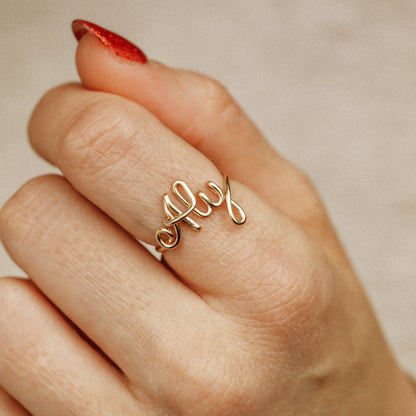 Personalized gold wire ring with the word "Aly" in script, worn on a hand with red nails.