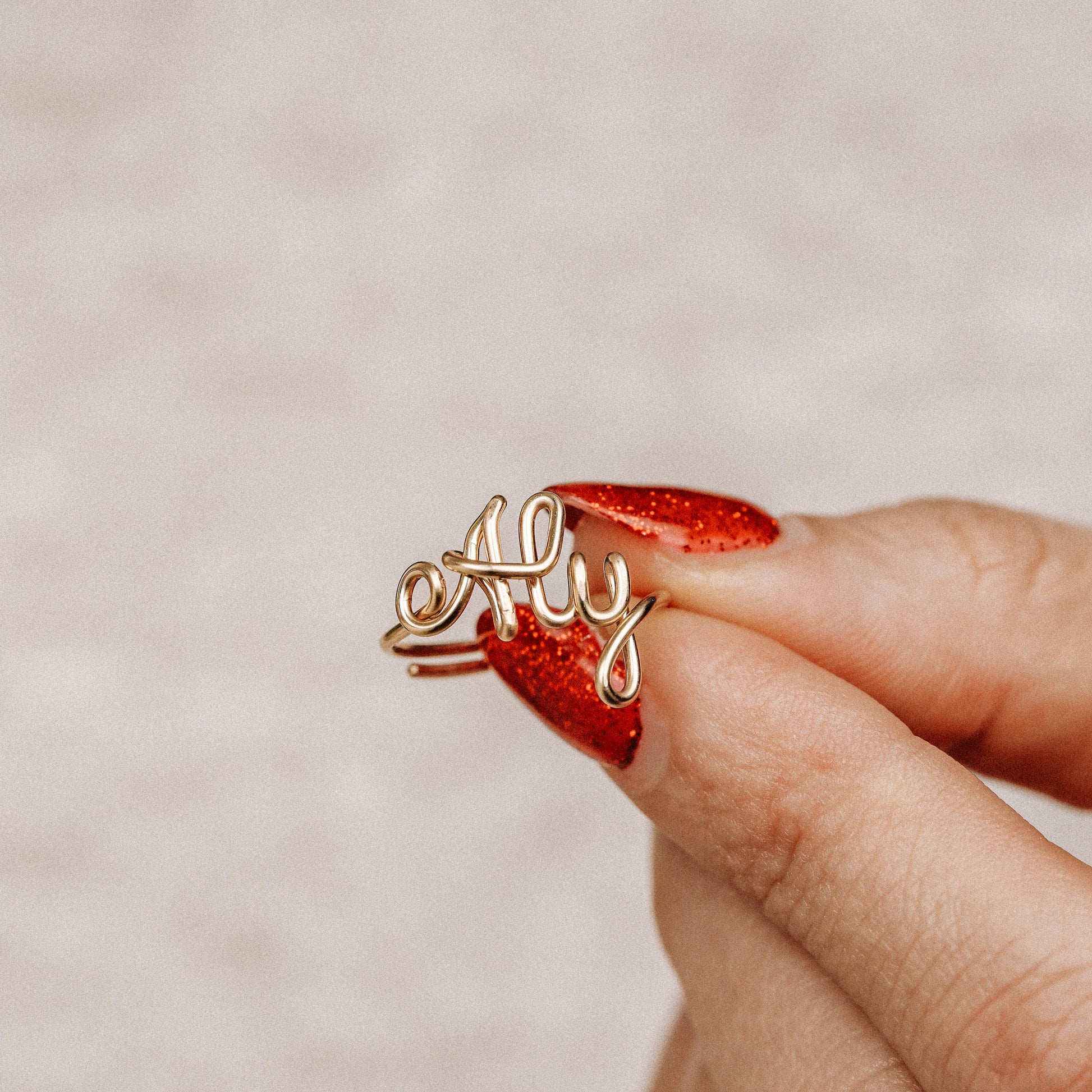 Personalized gold wire ring with the word "Aly" in script, worn on a hand with red nails.