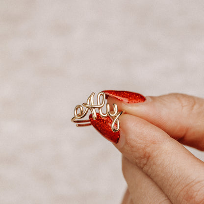Personalized gold wire ring with the word "Aly" in script, worn on a hand with red nails.