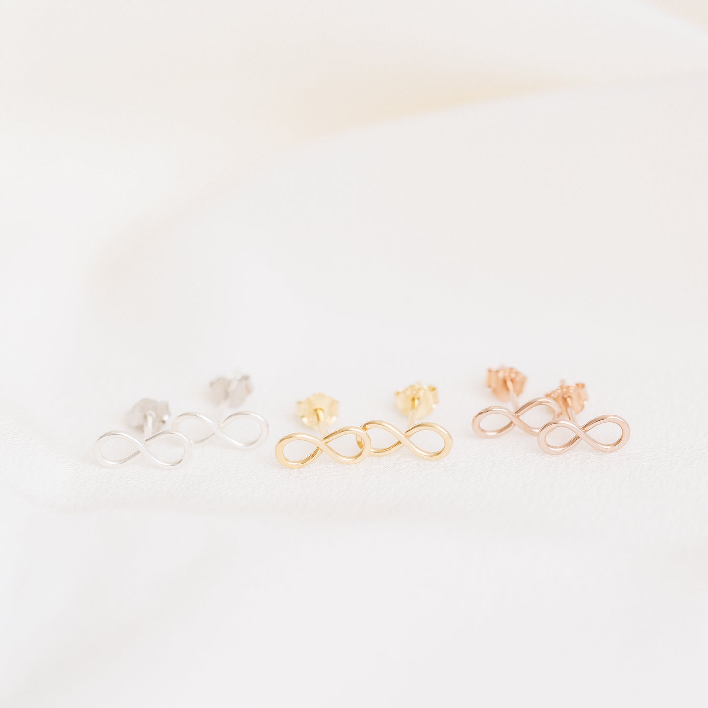 Tiny infinity stud earrings in silver gold and rose gold on soft white fabric