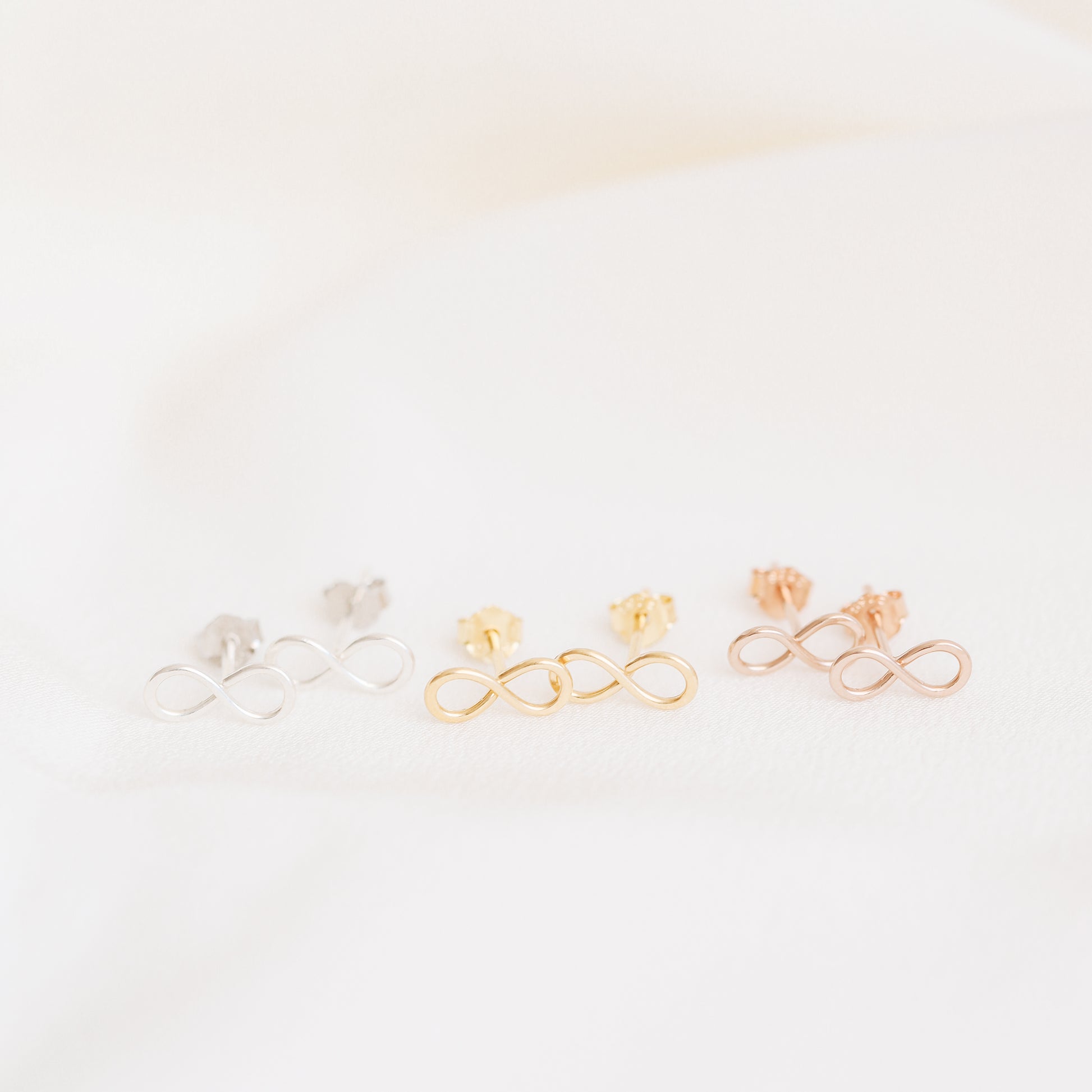 Tiny infinity stud earrings in silver gold and rose gold on soft white fabric