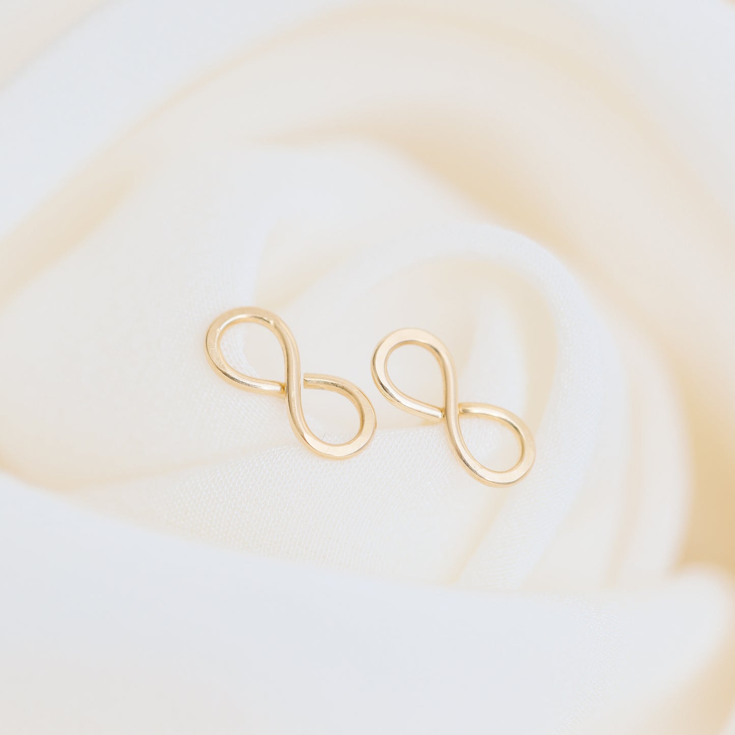 Minimal gold infinity earrings styled on soft ivory fabric