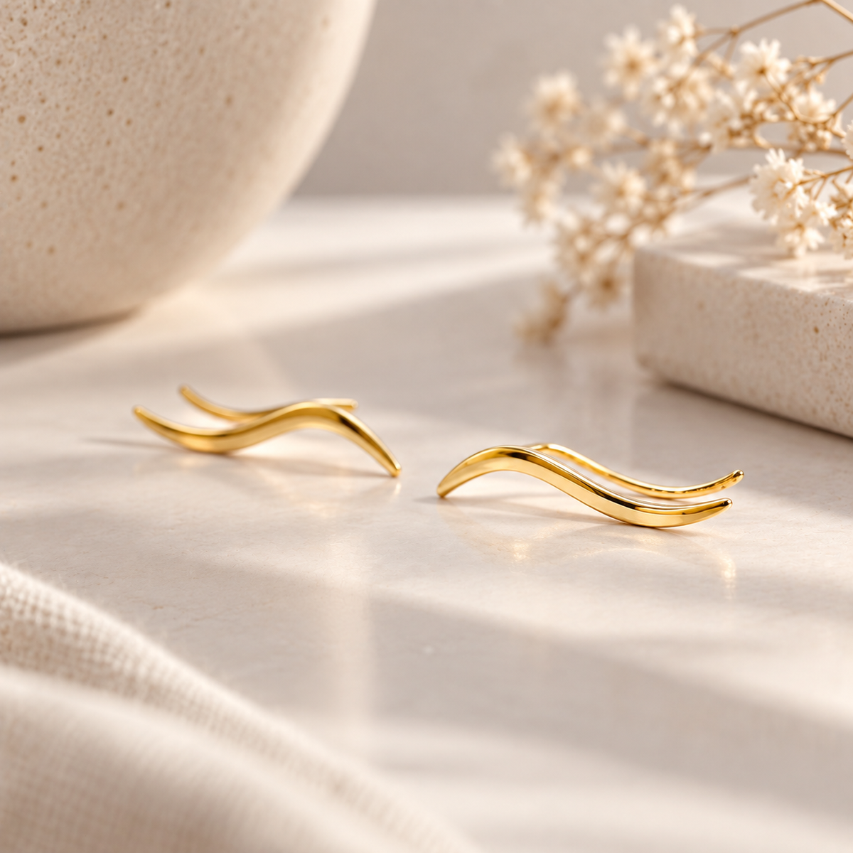 Pair of gold plated wave ear climbers displayed in soft natural light, featuring a flowing curved design with a sleek polished surface.