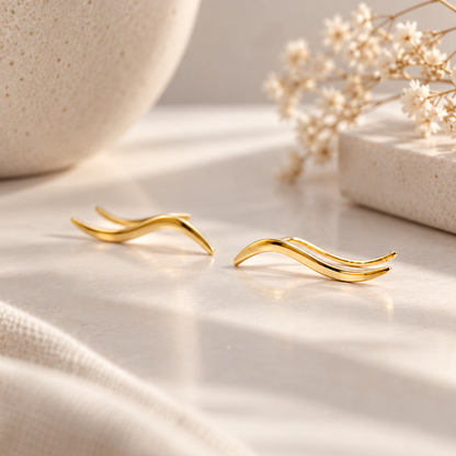 Pair of gold plated wave ear climbers displayed in soft natural light, featuring a flowing curved design with a sleek polished surface.