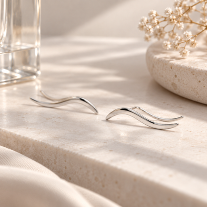 Matching left and right rhodium plated wave ear climbers displayed on a neutral stone surface, showing the mirrored curved silhouettes and high-polish finish.