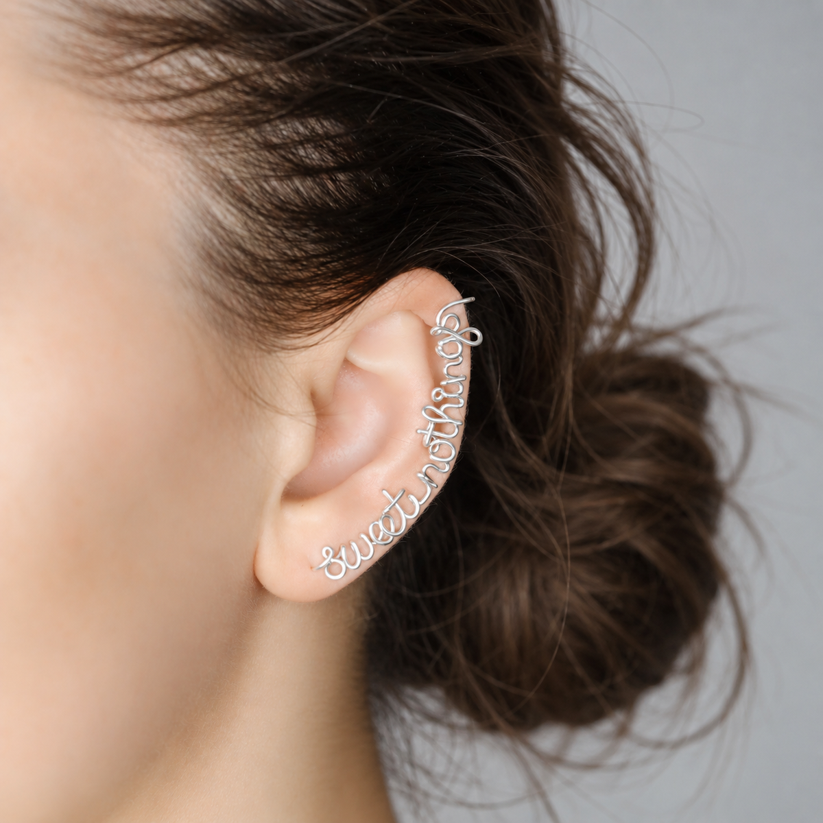 Personalized silver name ear climber handmade in flowing script, following the curve of the ear from the lobe upward, shown with a soft brown low bun hairstyle.