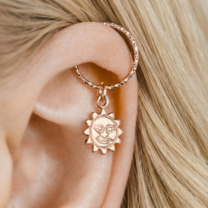 Close-up of a 18k rose gold endless hoop earring with a smiling sun charm pendant on a person's helix piercing, diamond-cut texture.
