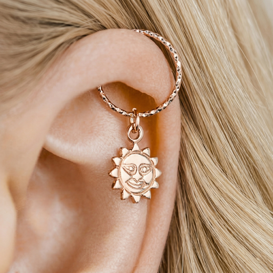 Close-up of a 18k rose gold endless hoop earring with a smiling sun charm pendant on a person's helix piercing, diamond-cut texture.