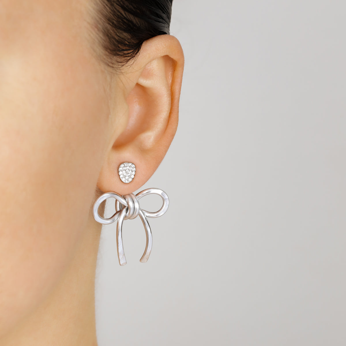 Silver medium bow ear jacket worn behind the earlobe with a sparkling stud, highlighting the lightweight hammered aluminum design.