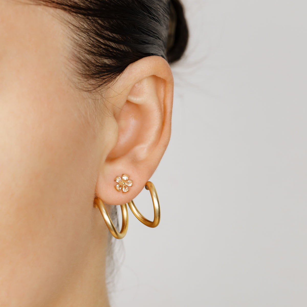 Gold double loop Ear Jacket shown worn in two different orientations behind the earlobe with a small floral stud, demonstrating how the curved design can change position around the ear.