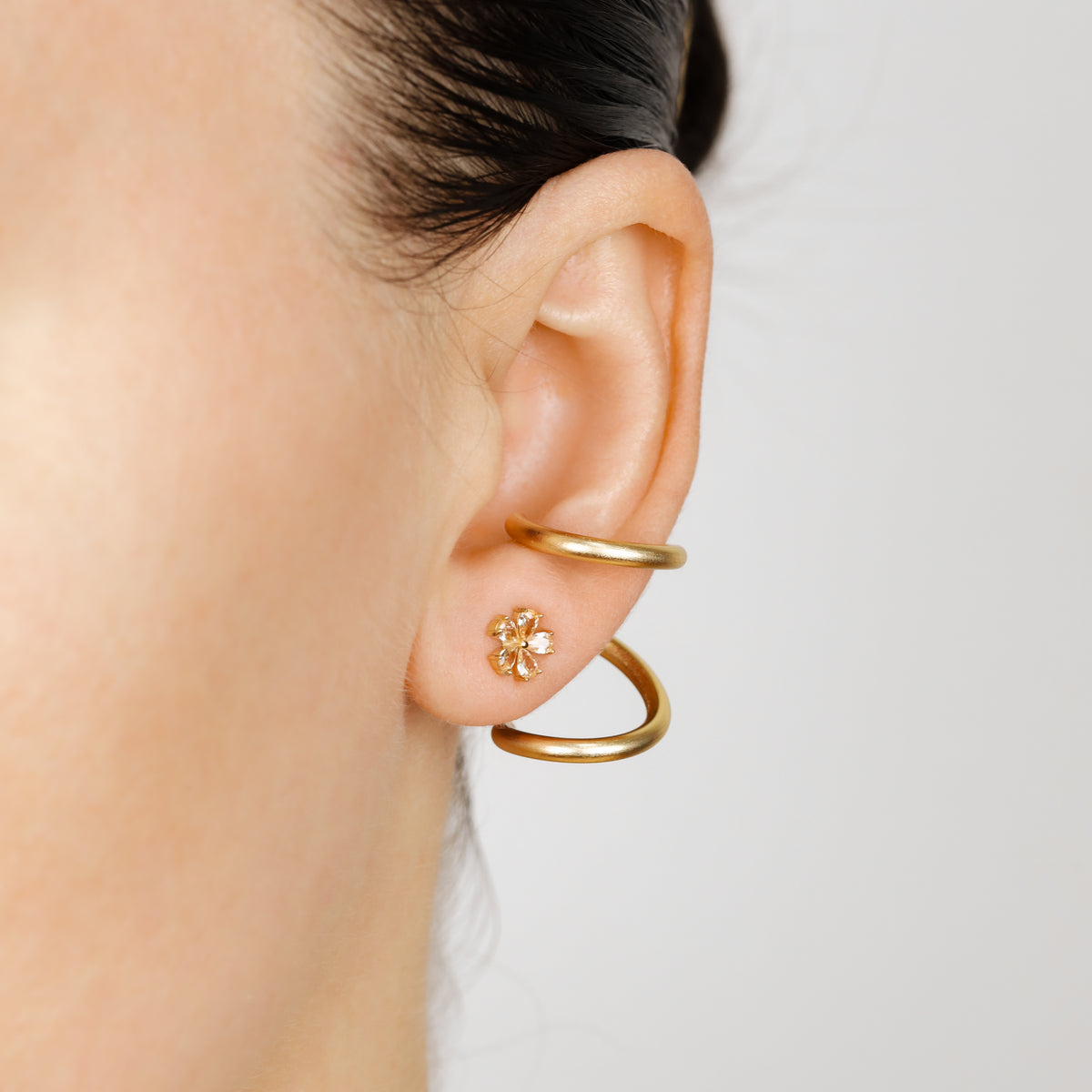 Pair of gold wavy double loop Ear Jackets worn behind the earlobe with a floral stud, showing the sculptural curved shape framing the lower part of the ear.