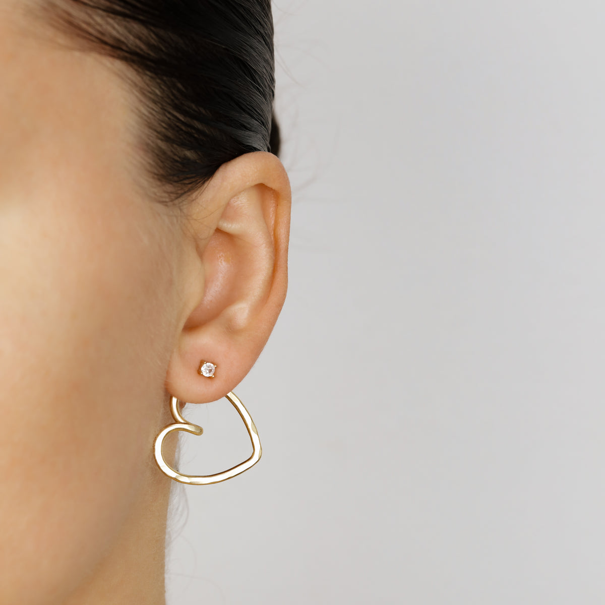 Gold open heart Ear Jacket worn behind the earlobe, showing how the lightweight hammered heart shape sits beneath the visible front stud.