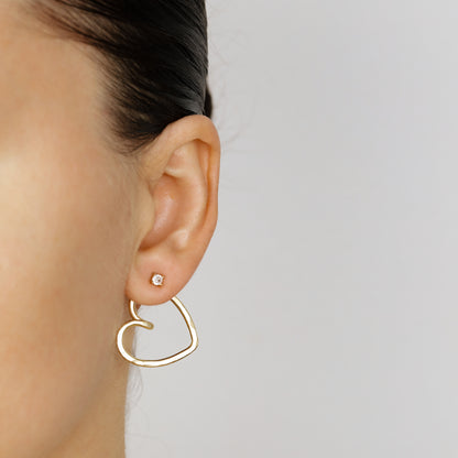 Gold open heart Ear Jacket worn behind the earlobe, showing how the lightweight hammered heart shape sits beneath the visible front stud.