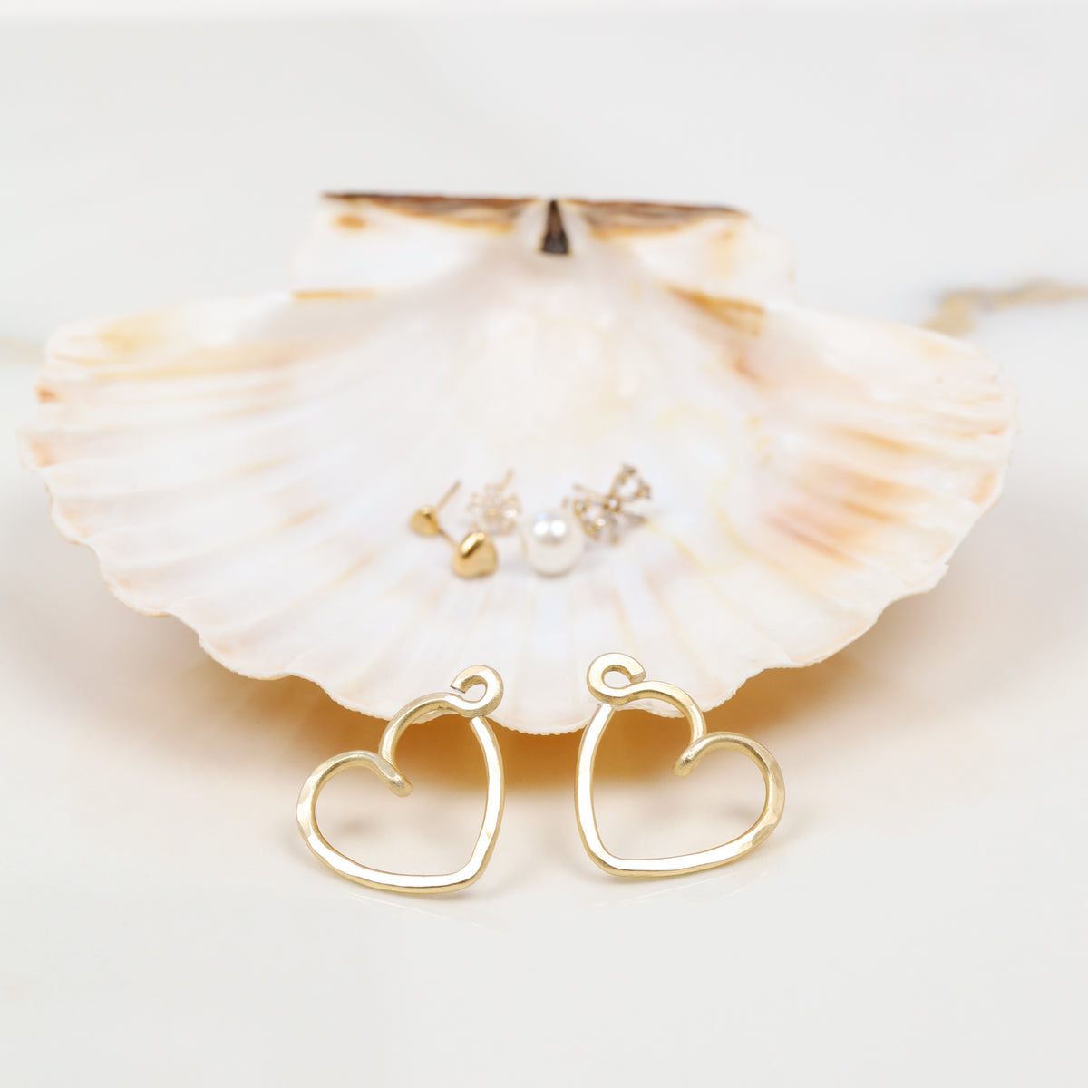 Pair of handmade gold heart Ear Jackets displayed in front of a natural shell with assorted stud earrings shown in the background as styling examples.