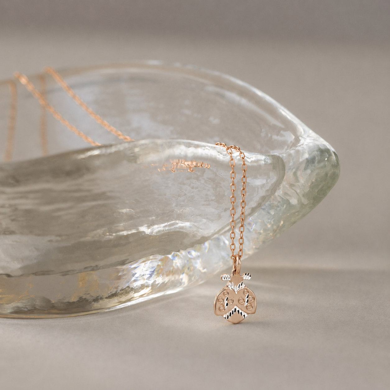 Gold ladybug necklace displayed beside textured glass bowl with soft neutral background