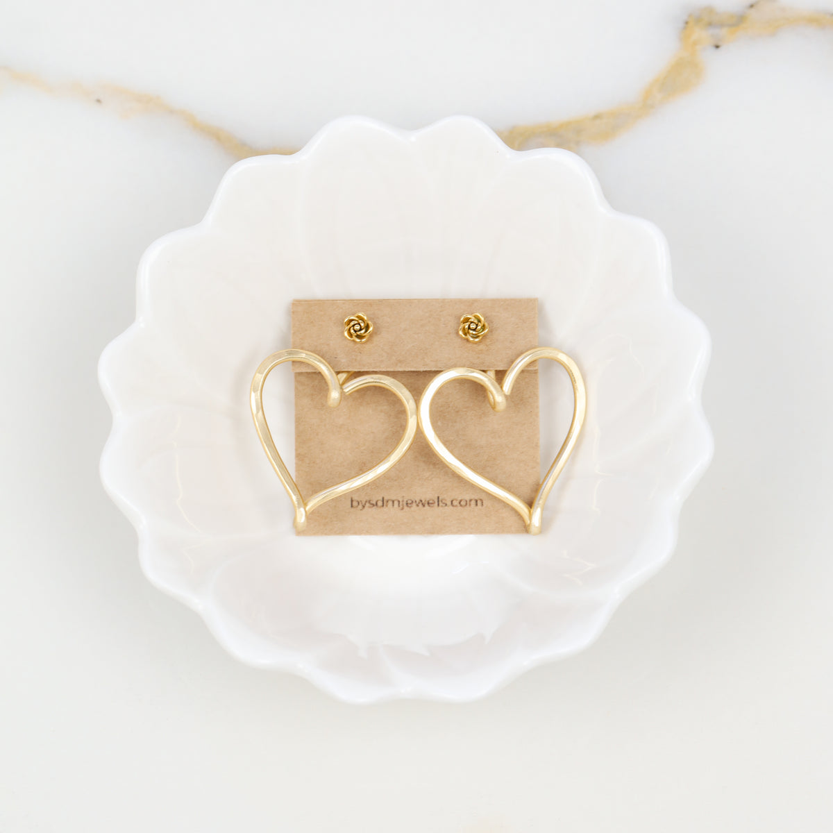 Pair of gold hammered Large Heart Ear Jackets displayed on a jewelry card inside a white dish with small rose-shaped stud earrings as a styling example.