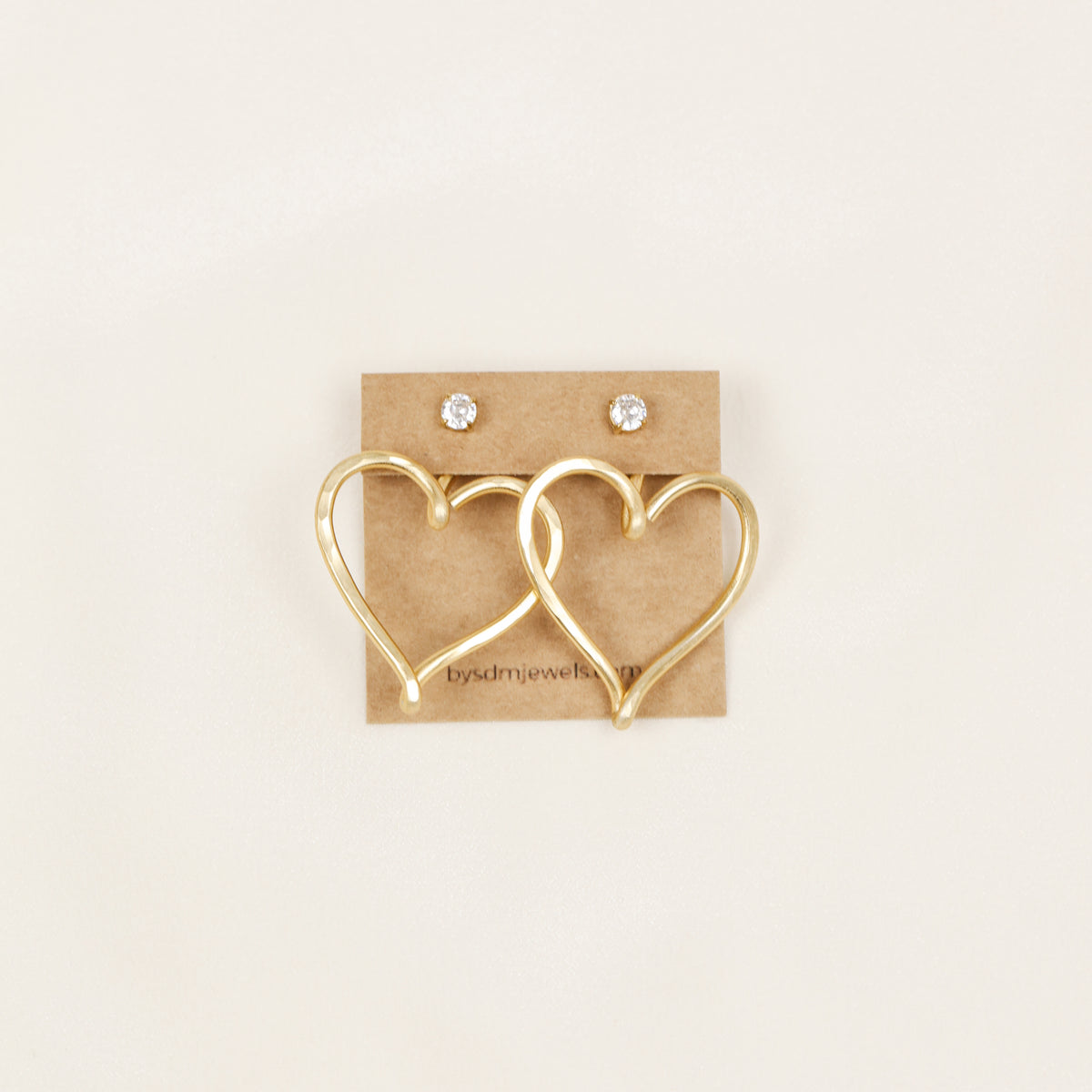 Pair of gold hammered Large Heart Ear Jackets displayed on a jewelry card inside a white dish with small rose-shaped stud earrings as a styling example.