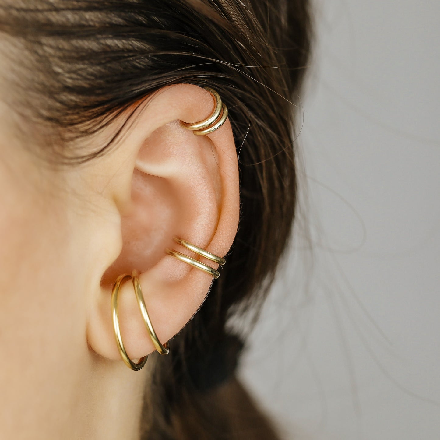 Thick smooth no-piercing ear cuff shown worn on different areas of the ear, including lobe, conch, cartilage and upper helix, with a rounded wrap design available in sterling silver, 18k yellow gold plated sterling silver, 18k rose gold plated sterling silver and black aluminum.