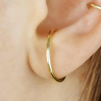 Thick smooth no-piercing ear cuff shown worn on different areas of the ear, including lobe, conch, cartilage and upper helix, with a rounded wrap design available in sterling silver, 18k yellow gold plated sterling silver, 18k rose gold plated sterling silver and black aluminum.