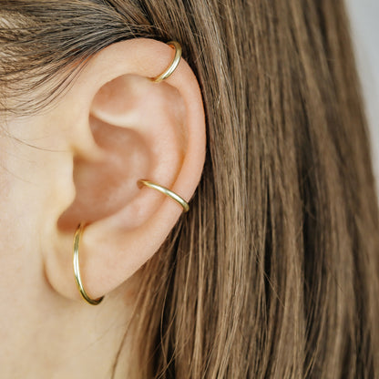Thick smooth no-piercing ear cuff shown worn on different areas of the ear, including lobe, conch, cartilage and upper helix, with a rounded wrap design available in sterling silver, 18k yellow gold plated sterling silver, 18k rose gold plated sterling silver and black aluminum.