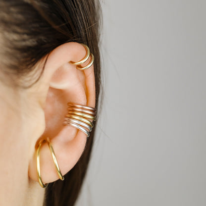 Thick smooth no-piercing ear cuff shown worn on different areas of the ear, including lobe, conch, cartilage and upper helix, with a rounded wrap design available in sterling silver, 18k yellow gold plated sterling silver, 18k rose gold plated sterling silver and black aluminum.