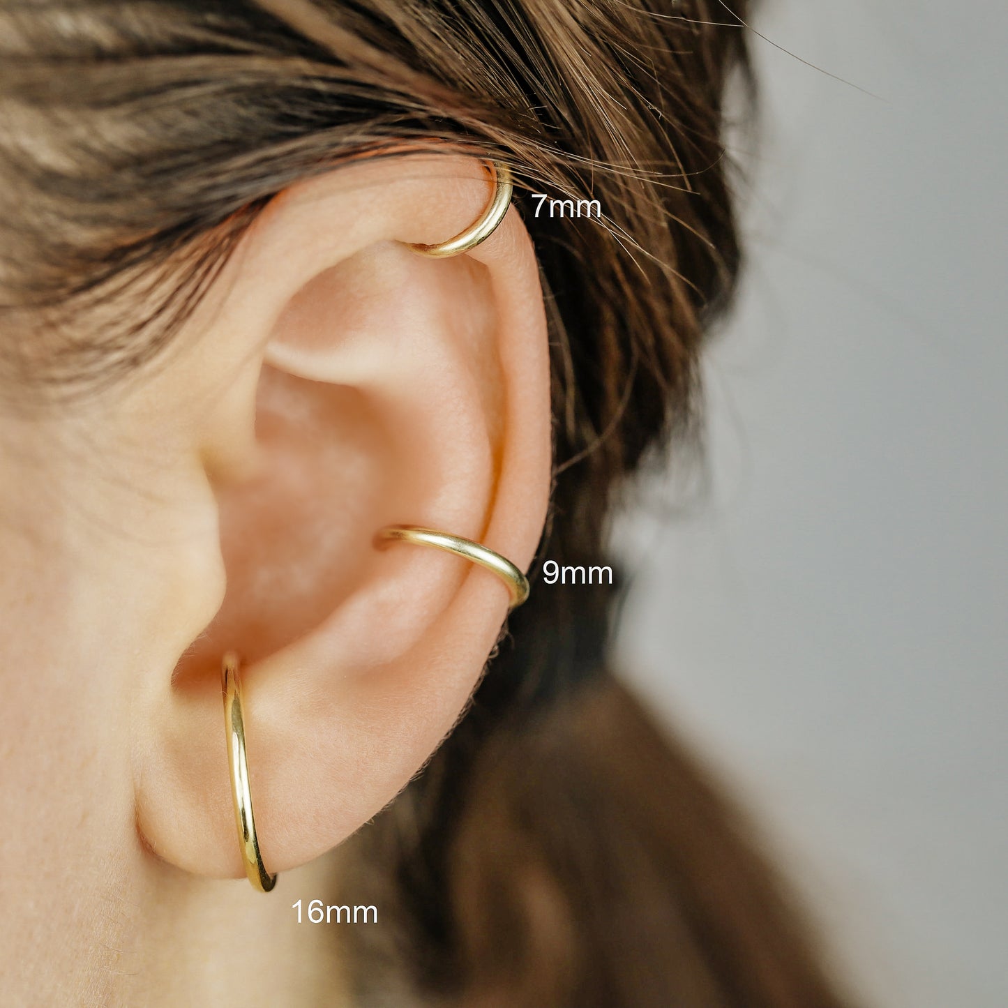 Thick smooth no-piercing ear cuff shown worn on different areas of the ear, including lobe, conch, cartilage and upper helix, with a rounded wrap design available in sterling silver, 18k yellow gold plated sterling silver, 18k rose gold plated sterling silver and black aluminum.