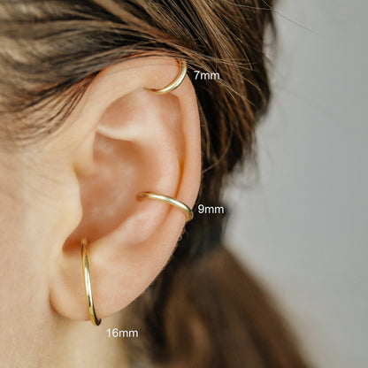 Thick smooth no-piercing ear cuff shown worn on different areas of the ear, including lobe, conch, cartilage and upper helix, with a rounded wrap design available in sterling silver, 18k yellow gold plated sterling silver, 18k rose gold plated sterling silver and black aluminum.