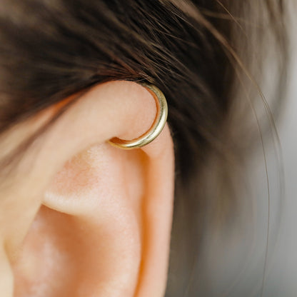 Thick smooth no-piercing ear cuff shown worn on different areas of the ear, including lobe, conch, cartilage and upper helix, with a rounded wrap design available in sterling silver, 18k yellow gold plated sterling silver, 18k rose gold plated sterling silver and black aluminum.