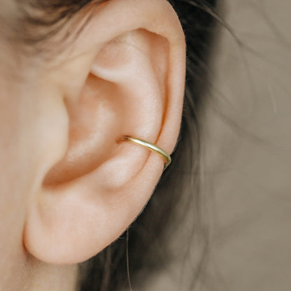 Thick smooth no-piercing ear cuff shown worn on different areas of the ear, including lobe, conch, cartilage and upper helix, with a rounded wrap design available in sterling silver, 18k yellow gold plated sterling silver, 18k rose gold plated sterling silver and black aluminum.